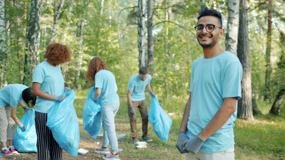 Portrait of Arabian eco-activist wearing volunteer t-shirt and gloves in forest smiling looking at camera while youth collecting junk in bin bags outside.