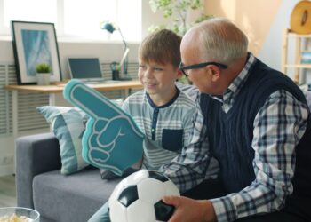 Joyful family grandfather and grandson are watching football on TV cheering hugging celebrating victory. Sports fans, modern lifestyle and leisure concept.