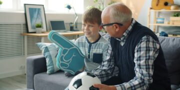 Joyful family grandfather and grandson are watching football on TV cheering hugging celebrating victory. Sports fans, modern lifestyle and leisure concept.