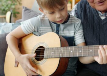 Elderly man grandfather is teaching grandson to play the guitar in apartment bonding enjoying home education. Musical instruments and childhood concept.