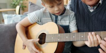 Elderly man grandfather is teaching grandson to play the guitar in apartment bonding enjoying home education. Musical instruments and childhood concept.