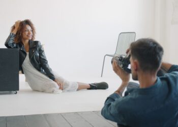 Sexy girl in fashionable clothing is posing for camera in studio working with professional photographer sitting on floor on white background. People and lifestyle concept.