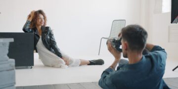 Sexy girl in fashionable clothing is posing for camera in studio working with professional photographer sitting on floor on white background. People and lifestyle concept.