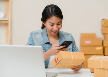 Beautiful smart Asian young entrepreneur business woman owner of SME checking product on stock scan qr code working at home. Small business owner at home office concept.