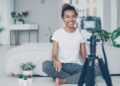 Female blogger is recording video for videolog looking at camera, smiling and talking emotionally sitting on bedroom floor at home. People, technology and social media concept.