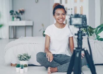 Female blogger is recording video for videolog looking at camera, smiling and talking emotionally sitting on bedroom floor at home. People, technology and social media concept.