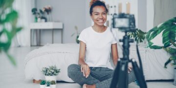 Female blogger is recording video for videolog looking at camera, smiling and talking emotionally sitting on bedroom floor at home. People, technology and social media concept.